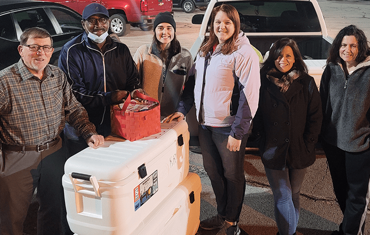 Six people stand together outside at night, smiling at the camera. They are gathered around two large white coolers and a red basket, with parked vehicles visible in the background. Kinsman Redeemer Homeless Ministry in Dickson County Tennessee