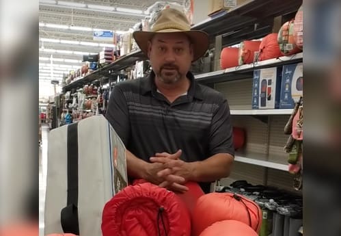 A man wearing a wide-brim hat and striped polo shirt stands in a store aisle with camping gear, including sleeping bags and a folding chair, displayed in front of him. Shelves of outdoor equipment line the aisle. Kinsman Redeemer Homeless Ministry in Dickson County Tennessee